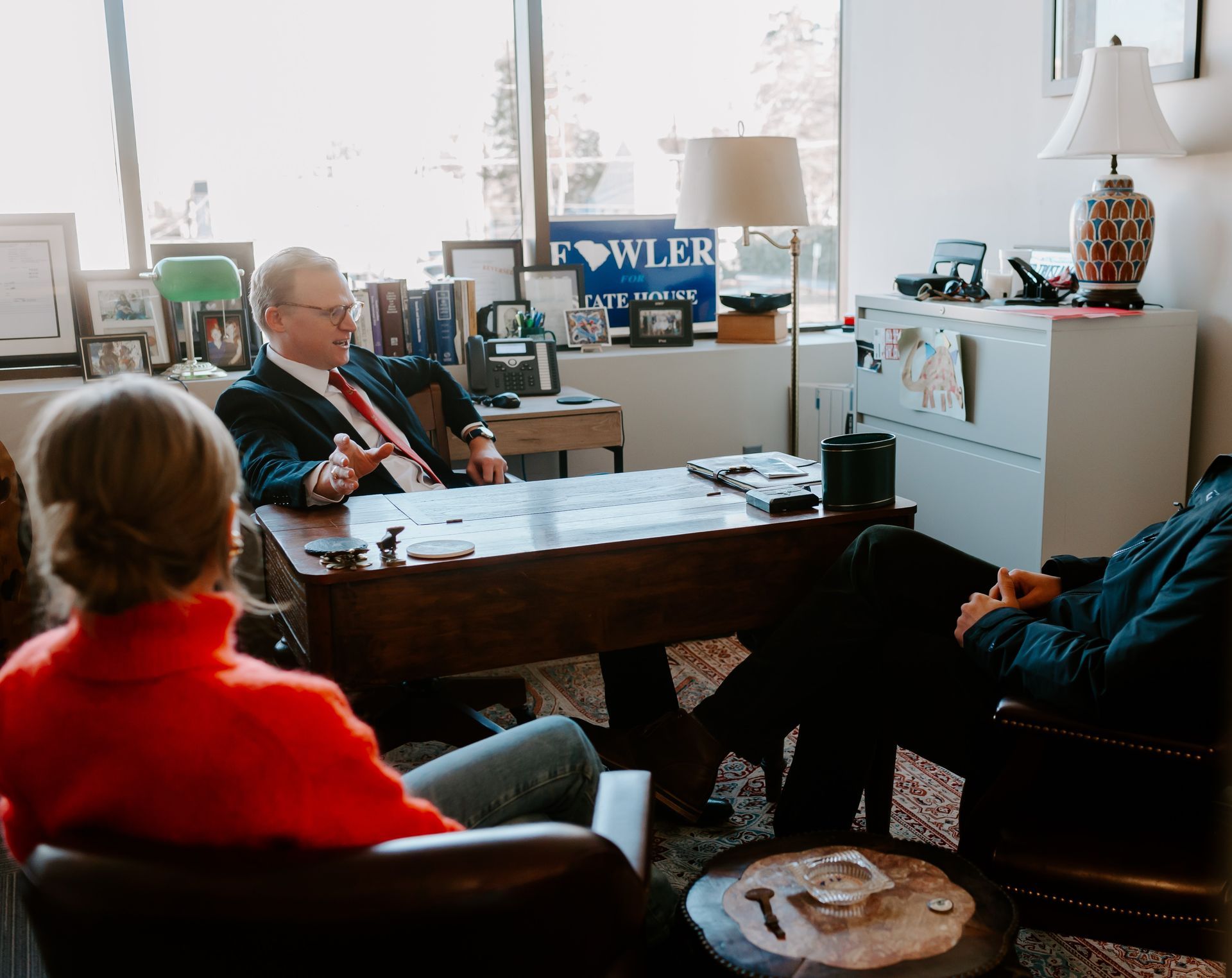 Brann Fowler in a suit sitting at a desk in an office, speaking with two other people