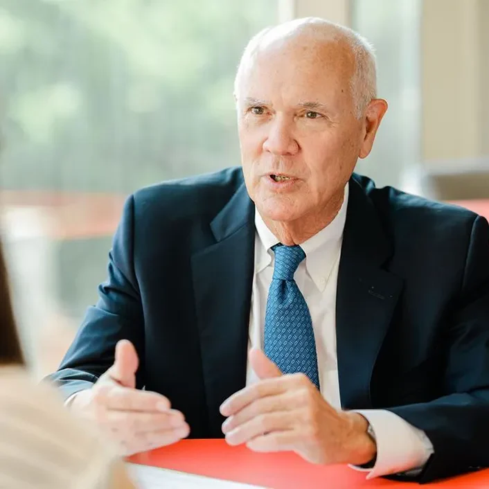 Joe Watson in a dark suit and blue tie engaged in a discussion, gesturing with hands across a table with blurred background.