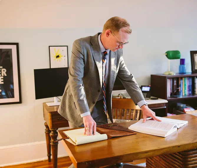 Brann Fowler in a suit leaning over a wooden desk, reviewing documents in an office with bookshelves and framed art.