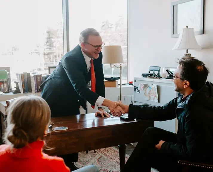 An older professional man in a suit shakes hands with a seated man across a wooden desk in a bright office.