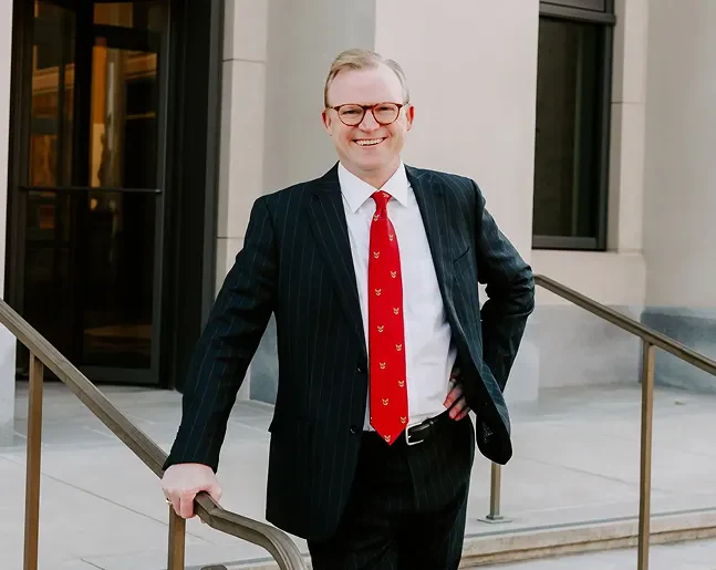 Brann Fowler in a black pinstripe suit with a red tie, standing outside a building holding a railing with one hand and the other on his hip.